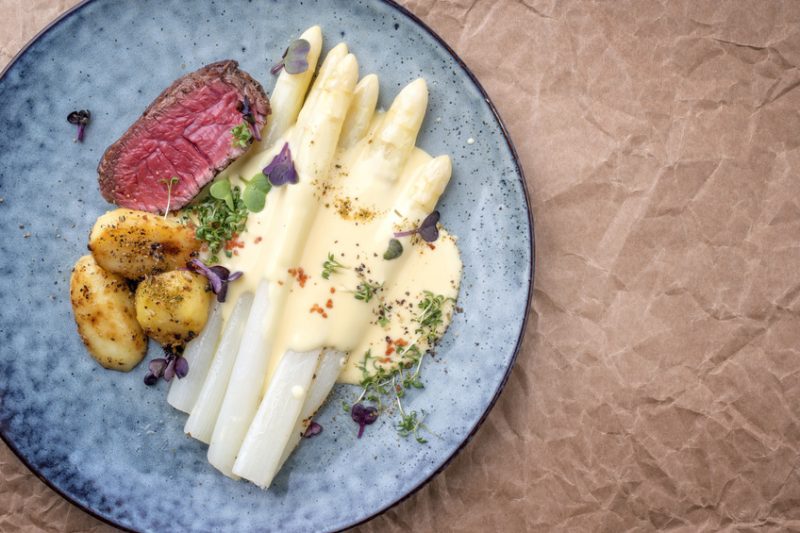 Modern barbecue dry aged sliced fillet steak with white asparagus in hollandaise sauce and roast potatoes as top view on a plate with copy space right