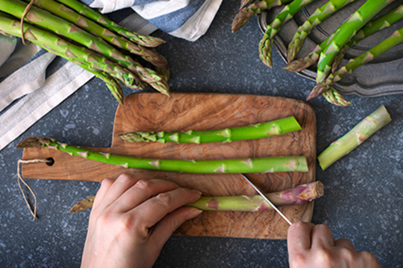 Fresh  raw asparagus on stone background. Women female hands cut asparagus on wooden cooking board. Top view