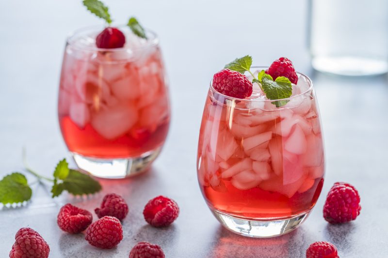 Two refreshing red translucent drinks with ice and raspberries. The raspberry juice is garnished with a green leaf, and there are pieces of ice and raspberries scattered around the drinks.