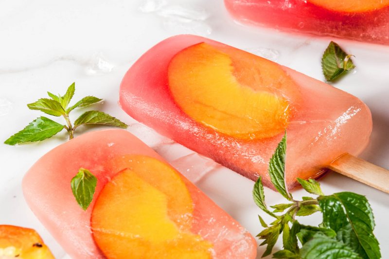 Summer desserts. Frozen drinks. Sweet fruit popsicles from frozen peach tea with mint. On a white marble table, with ingredients - peaches, mint, ice. Copy space