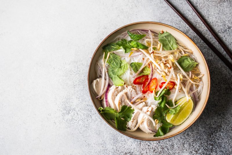 Traditional Vietnamese soup- pho ga in bowl with chicken and rice noodles, mint and cilantro, red onion, chili, bean sprouts and lime on grey background. Asian food. Copy space. Top view